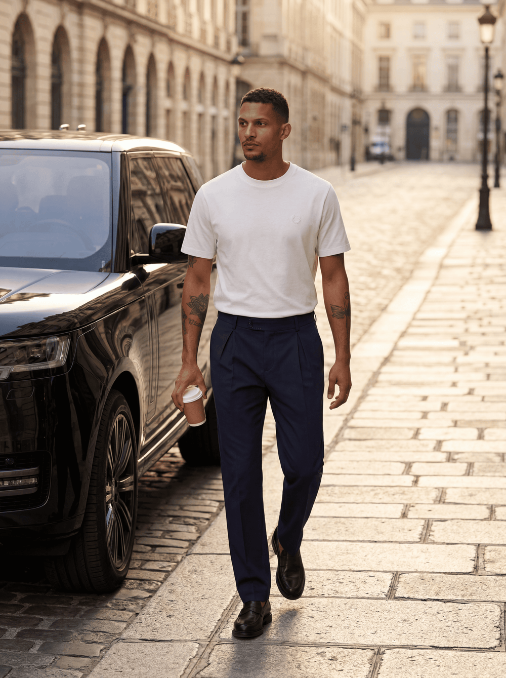 Man walking on a city street holding a coffee cup, with a black car and classical architecture in the background.